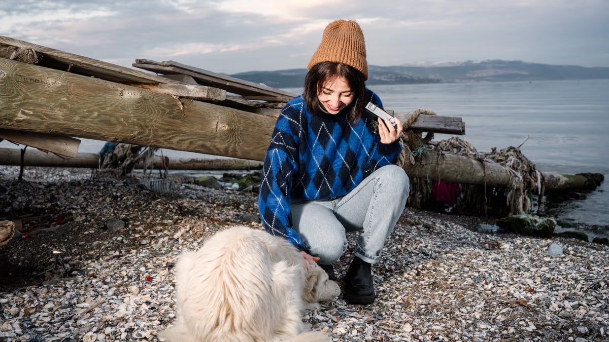 A young woman in a cozy sweater and beanie enjoys a serene moment by the beach with her dog.