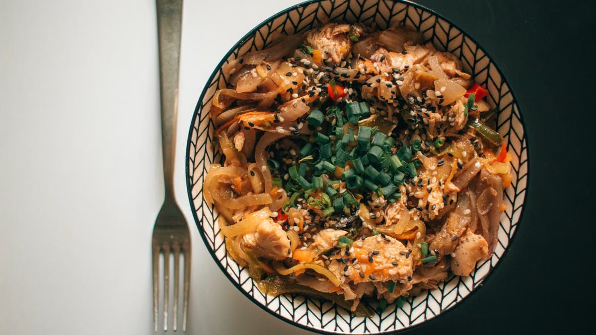 Close-up of a bowl of chicken stir-fry noodles with vegetables and sesame seeds, shot from above.